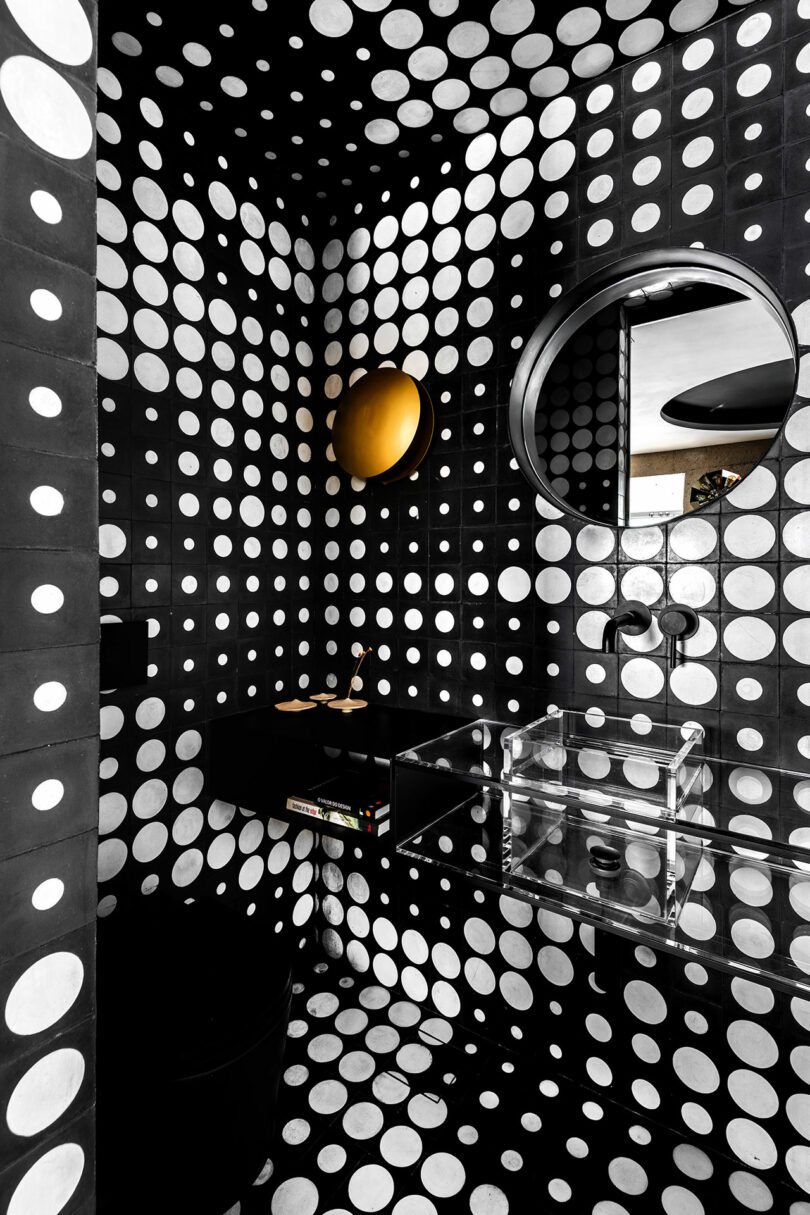 Black bathroom with white polka dot walls, a round black-framed mirror, a round wall light, a black sink, and a transparent rectangular basin.