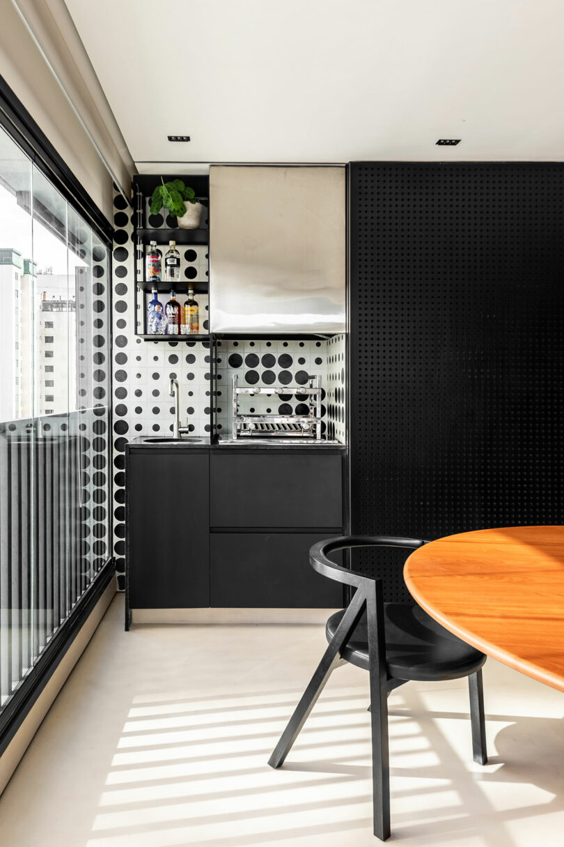 Modern kitchenette with black cabinets, polka dot backsplash, a small sink, and built-in grill; round wooden table and black chair in the foreground by large window.