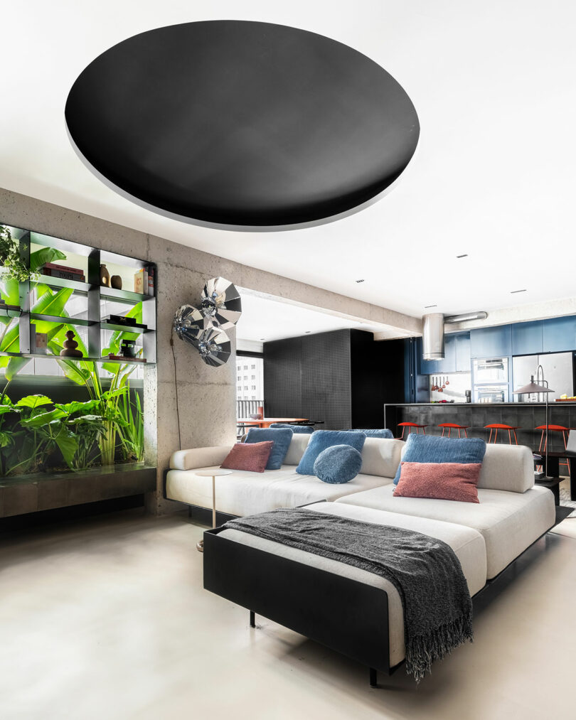 Modern living room with a white sectional sofa, colorful cushions, indoor plants, open kitchen with bar stools, and a large black circular ceiling fixture.