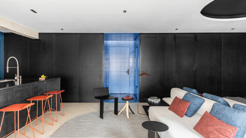 Modern living area with black walls, blue accents, a white sofa with blue cushions, red bar stools at a black kitchen counter, and a circular rug on a light floor.