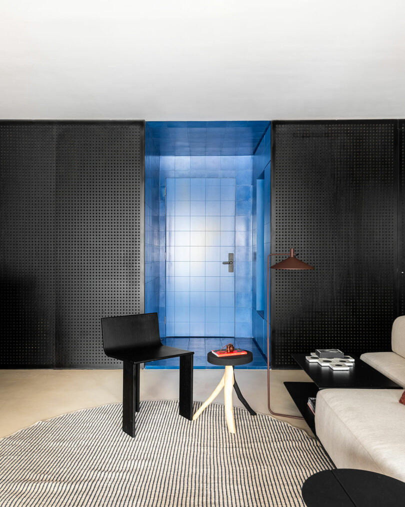 Modern living room with a black chair, a small light wood table, a beige sofa, and a striking blue tiled doorway framed by black perforated panels.