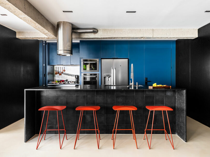 Modern kitchen with a black island, four red barstools, stainless steel appliances, blue cabinetry, and exposed concrete elements.