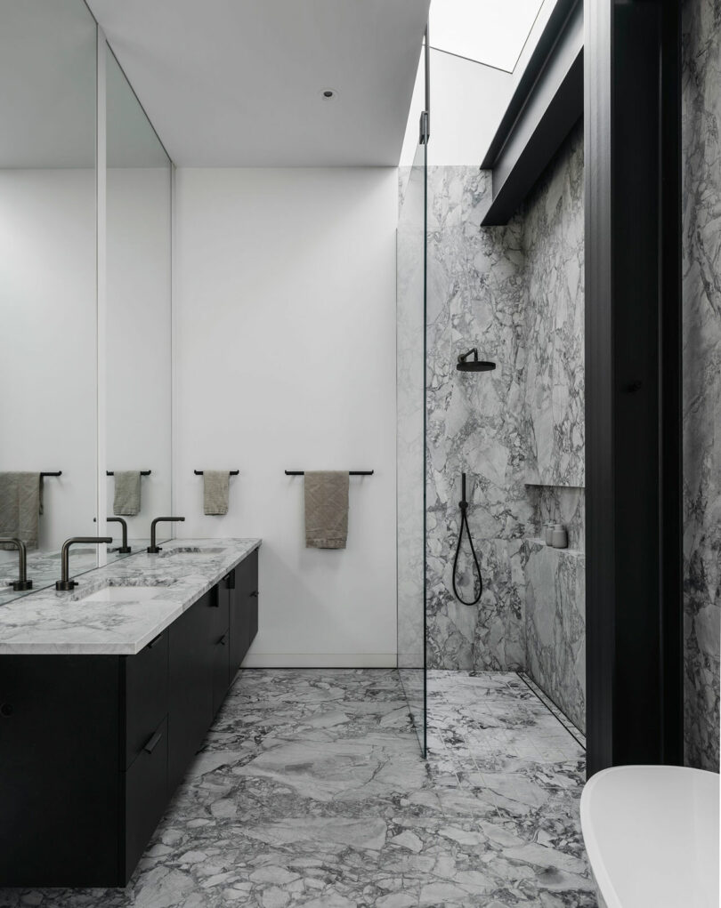 Modern bathroom with marble floors and walls, double sink vanity, glass-enclosed shower, black fixtures, and minimalistic decor.