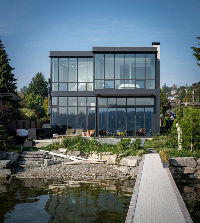 A modern two-story house with large glass windows stands by a lake, featuring a stone patio, dock, and surrounding greenery under a clear sky.