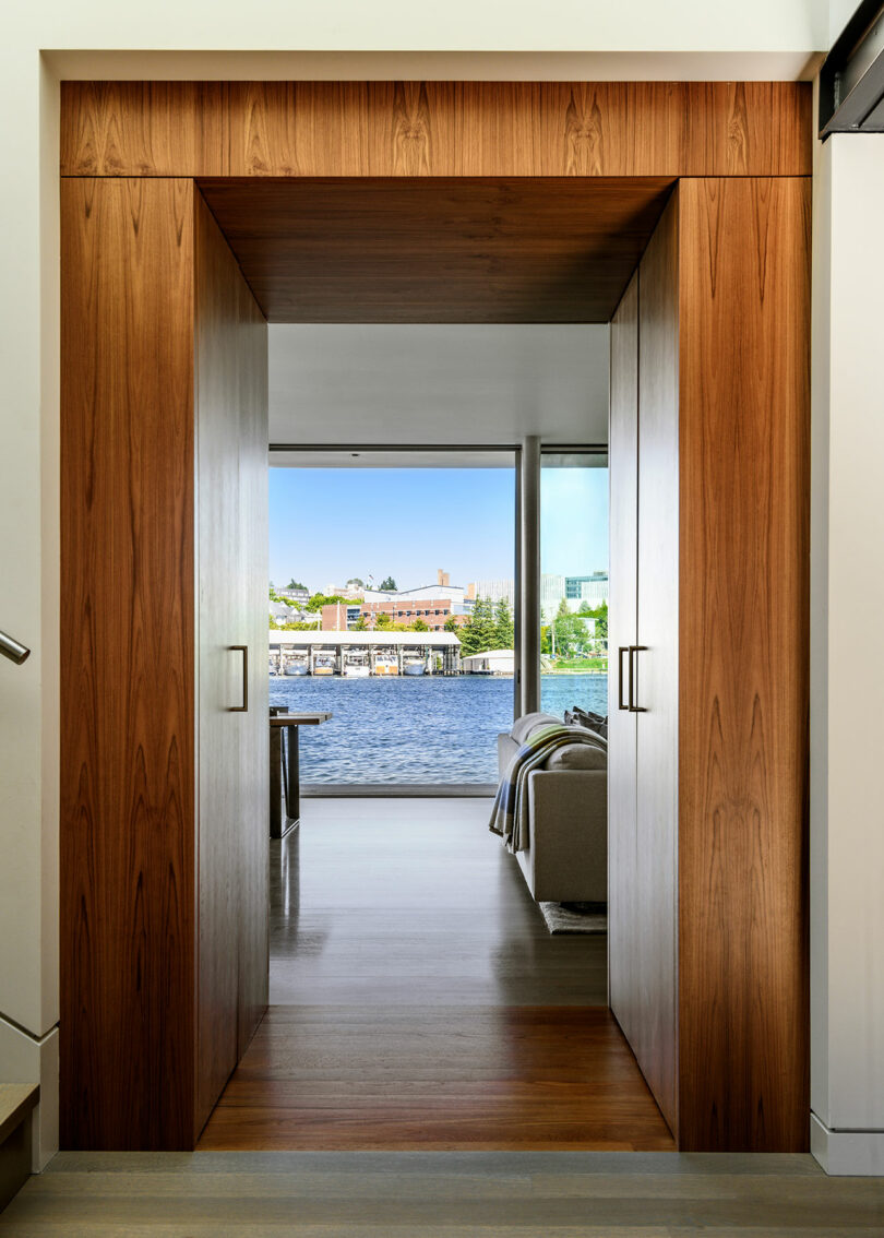 A wooden doorway frames a view of a modern living room with a sofa and large window overlooking water and buildings outside.