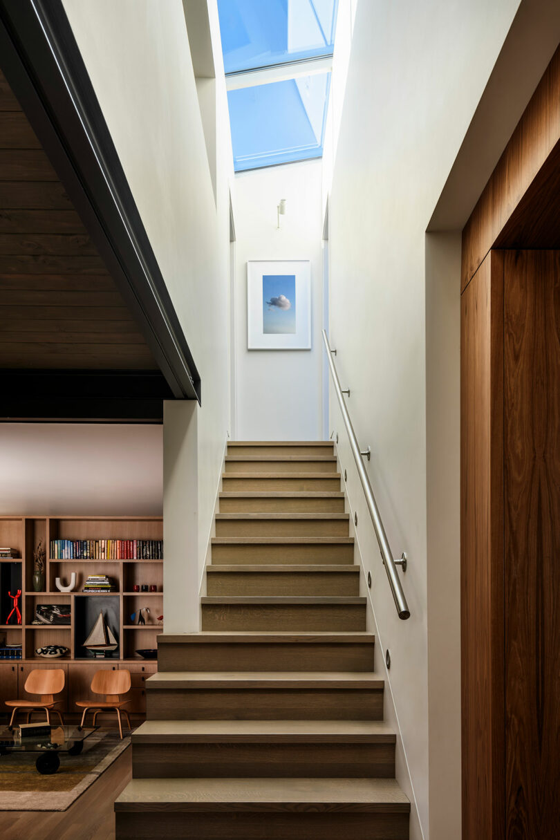 A modern wooden staircase with a metal handrail leads up to a skylight; a framed picture hangs on the wall, and a living room with shelves is visible to the left.