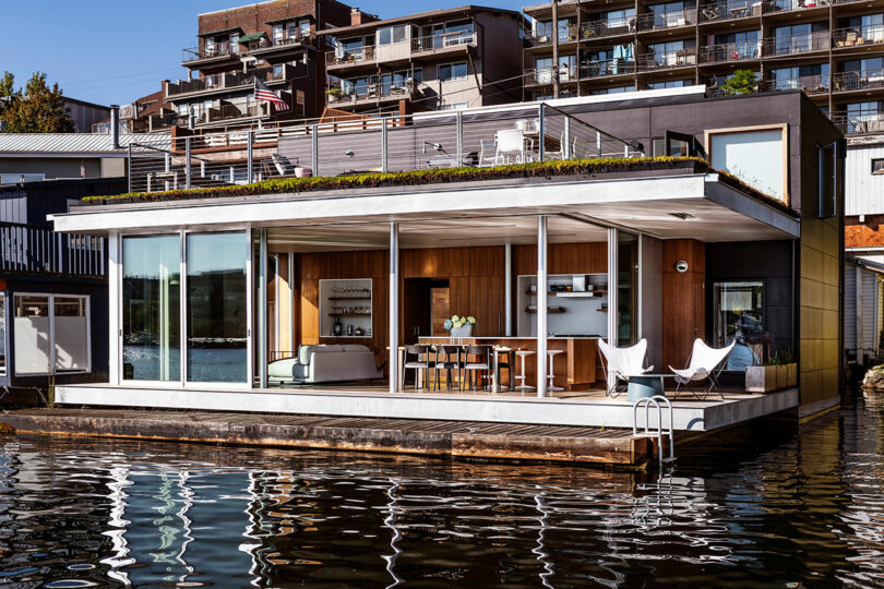 Modern houseboat with large glass windows, an open living and dining area, and a ladder leading into the water, docked in an urban setting with apartment buildings in the background.
