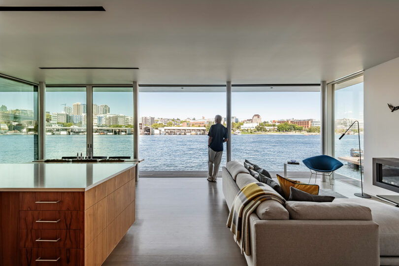 A person stands by large windows in a modern living room, overlooking a waterfront cityscape on a clear day.