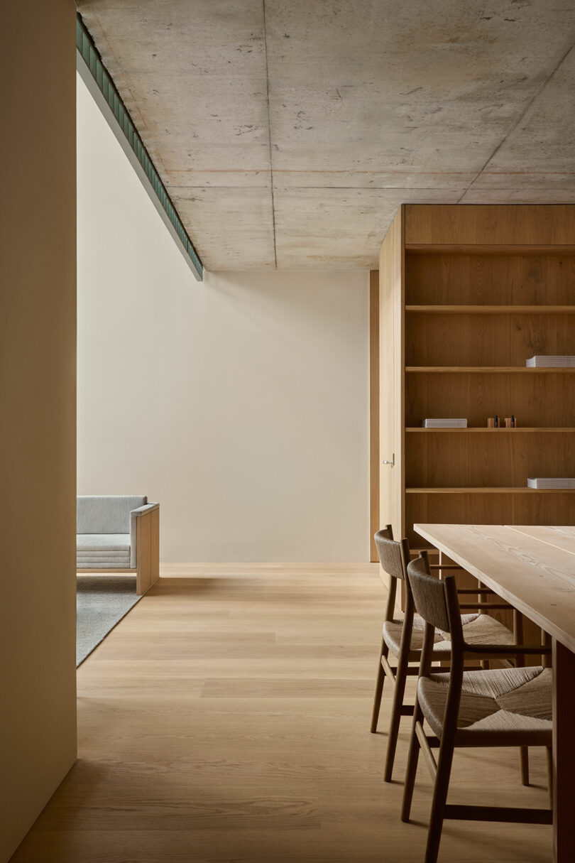 Minimalist dining room with wooden table and chairs, built-in wooden shelves, and large window allowing natural light. Concrete ceiling and light wood flooring.