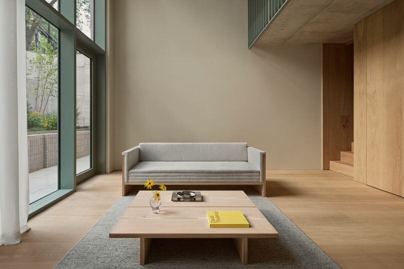 A minimalist living room with a light gray sofa, low wooden coffee table, small vase with yellow flowers, and large floor-to-ceiling windows.