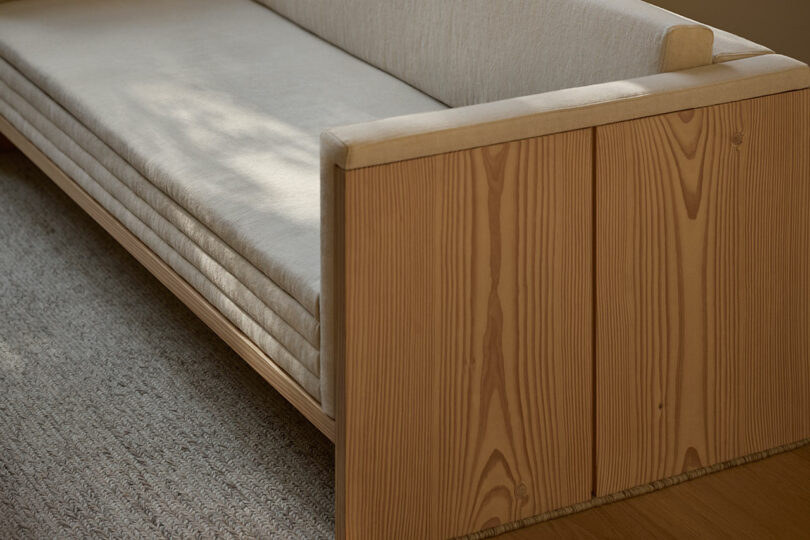 A close-up of a modern wooden sofa with light beige cushions, set on a textured rug and wooden floor.