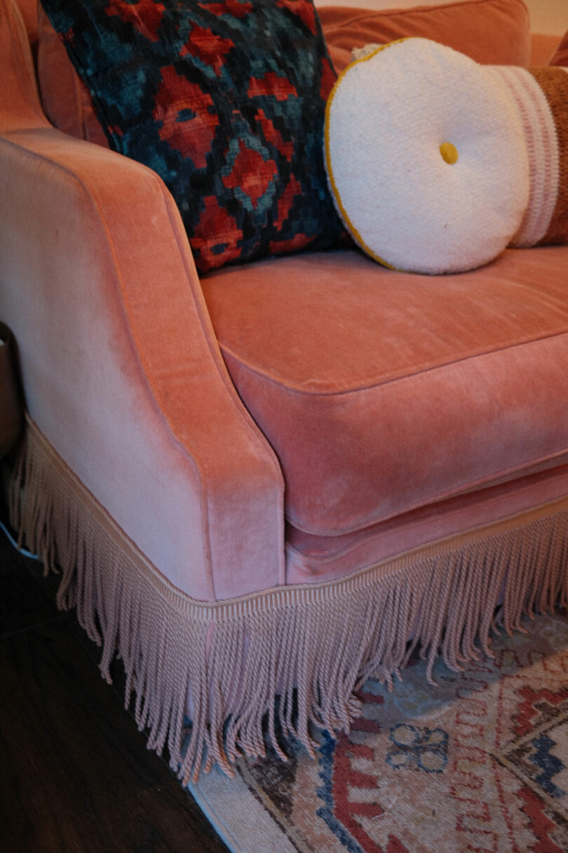 A pink velvet sofa with fringe along the bottom, decorated with a colorful patterned pillow and a round white pillow, adds a touch of Tom Parker-inspired flair. Part of a patterned rug is visible on the floor.