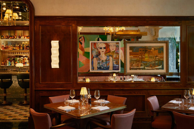 Cozy restaurant interior with set tables, leather chairs, wall art, and a view into a bar area with stools and bottles. Warm lighting and wood paneling create an inviting atmosphere.