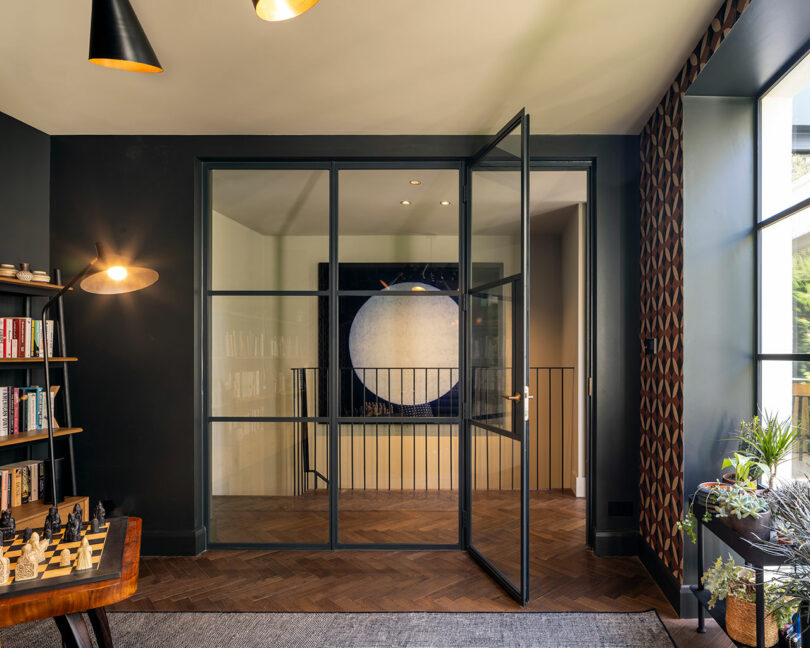 A modern room with dark walls, a glass door, a bookshelf, a chessboard table, potted plants, and a circular abstract artwork on the far wall.