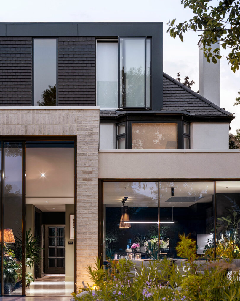 Modern two-story house with large glass windows, brick and wood exterior, and visible interior lighting at dusk, surrounded by plants and trees.
