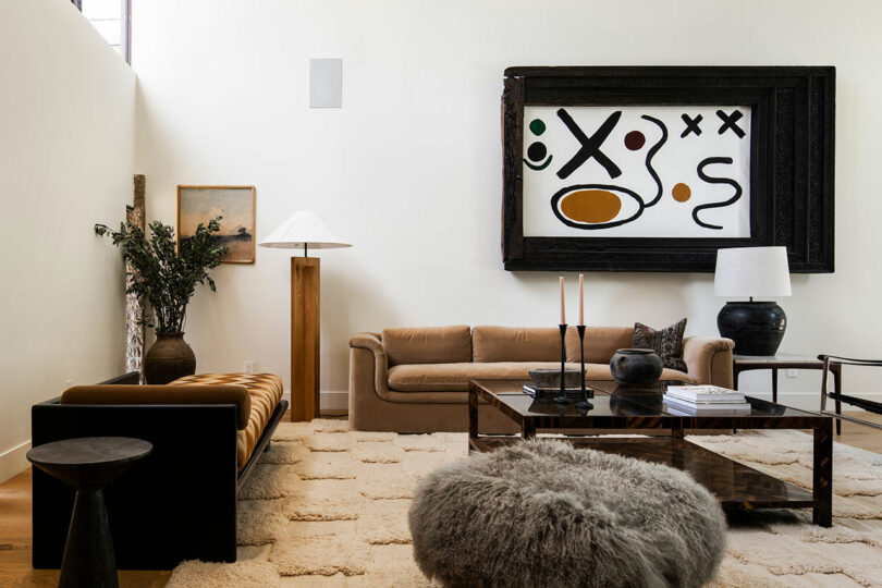Modern living room with two sofas, a large abstract painting, a glass coffee table, a floor lamp, a black table lamp, and decorative plants on a light patterned rug—perfect for your next Friday Five gathering.
