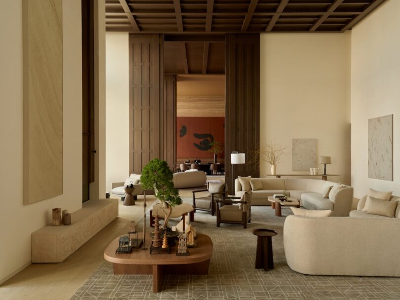 A spacious, modern living room with neutral tones reminiscent of skim milk, light beige furniture, wooden accents, large abstract art, and tall wooden doors opening to an adjoining room.