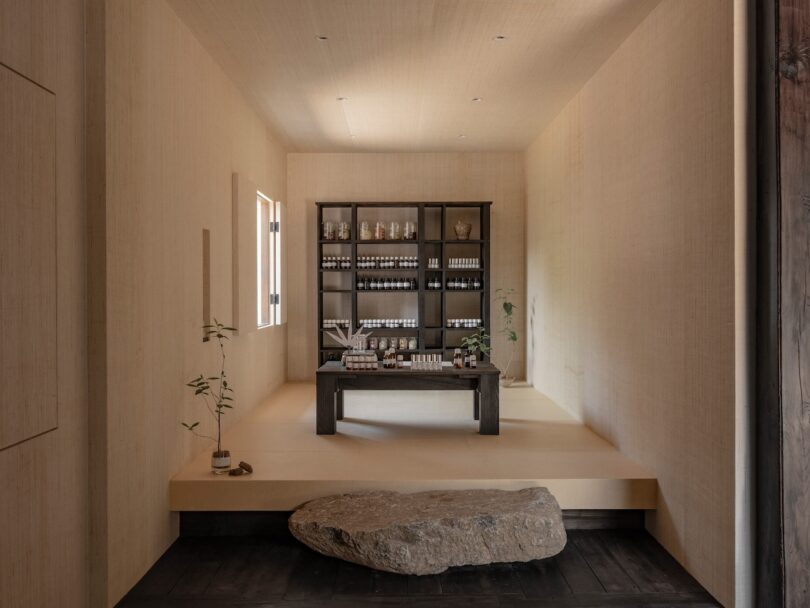 A minimalist room with light beige walls features a wooden table and shelves displaying neatly arranged bottles and jars of Skim Milk, with a stone step at the entrance and a small potted plant on the left.