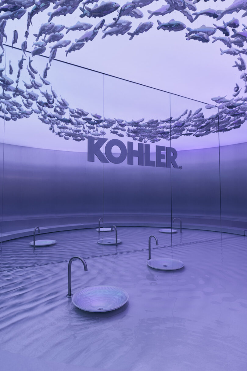 Modern showroom with four round sinks, metallic faucets, reflective walls with "Kohler" branding, and sculptural fish forms suspended from the ceiling, all bathed in purple light.