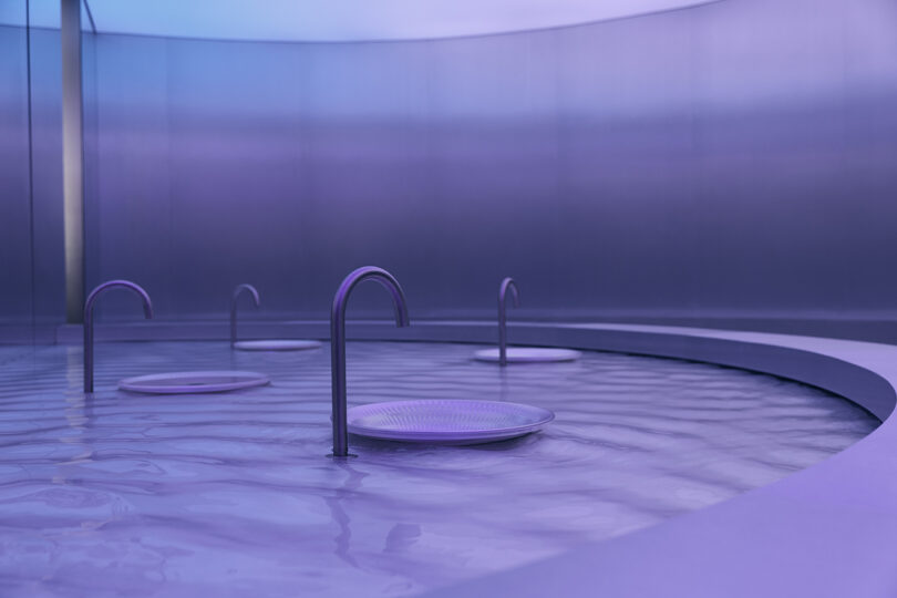 Circular pool with shallow water, featuring metal faucets and round plates on the surface, illuminated by purple-blue lighting.