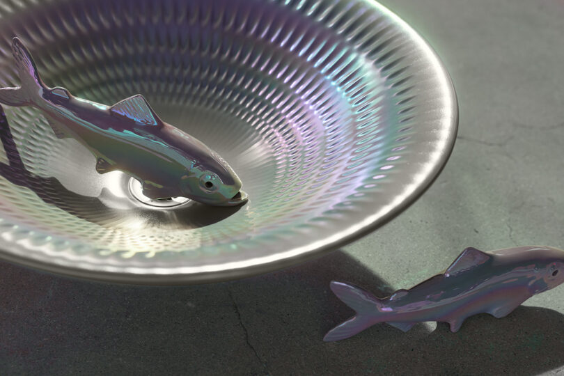 Two shiny, metallic fish models rest, one inside and one beside a textured, metallic bowl on a gray surface.