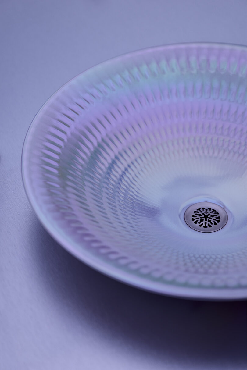 A close-up of a textured, iridescent plate with a circular pattern and a decorative drain-like design in the center, set against a light purple surface.