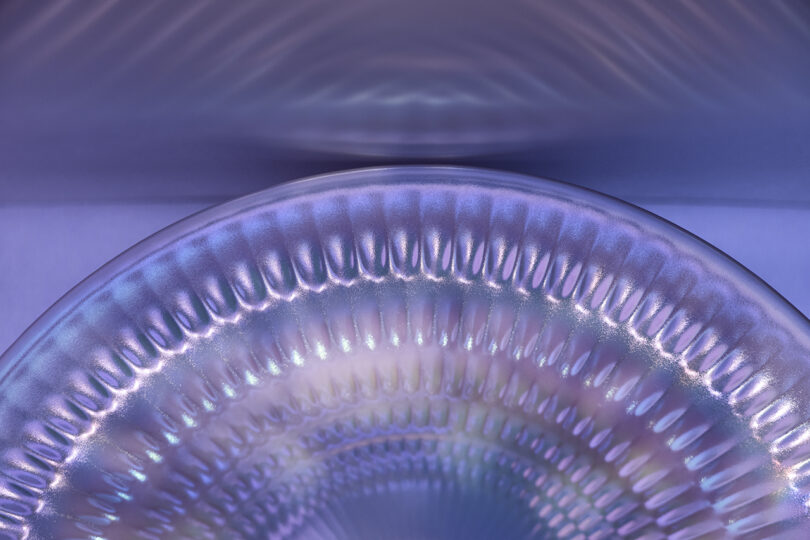 Close-up of a metallic, iridescent, ridged surface with a purple and blue color gradient, reflecting light in a wavy pattern.