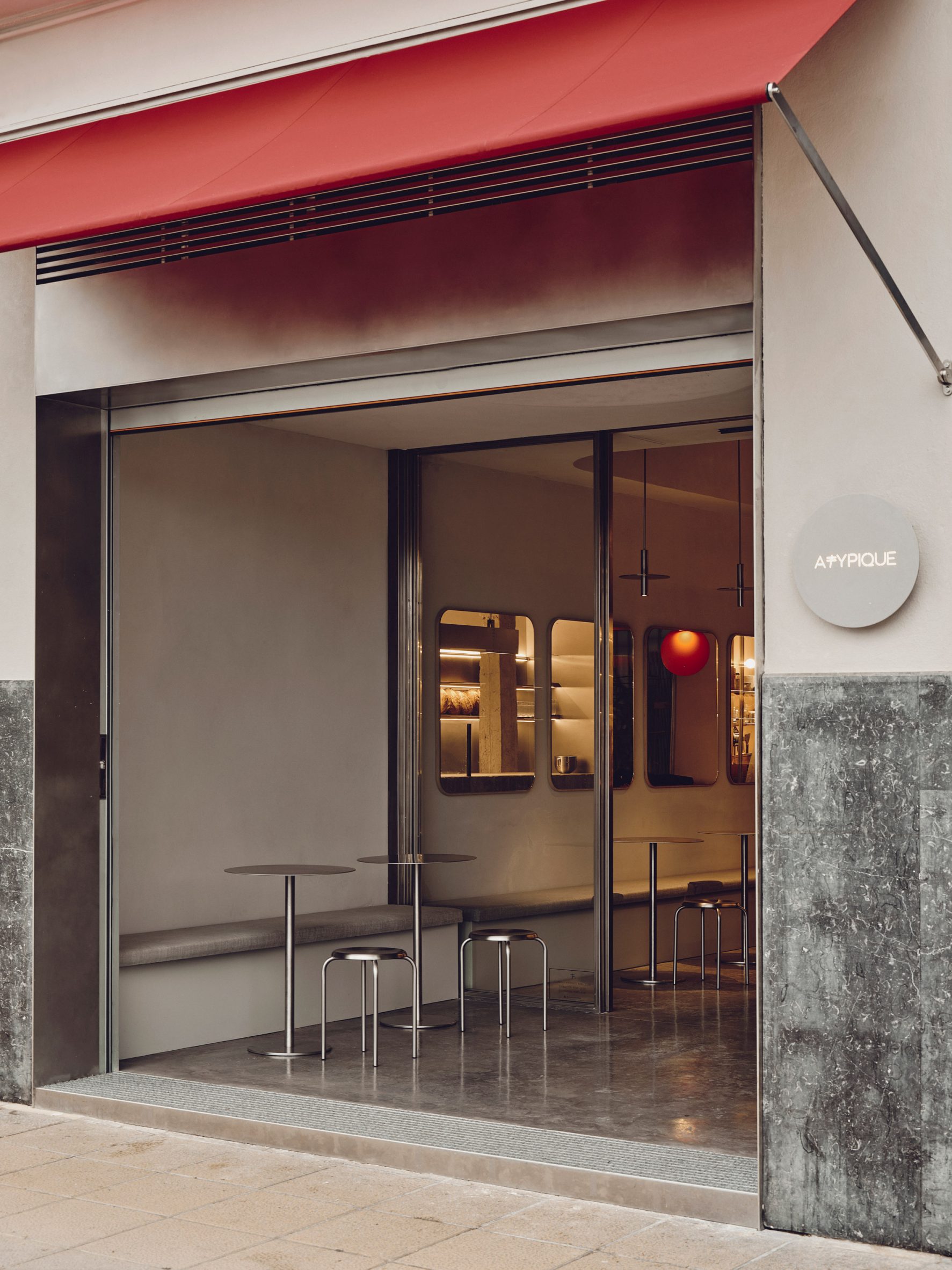 Modern café exterior with red awning, open glass façade, stainless-steel tables and stools, and minimalist interior view.
