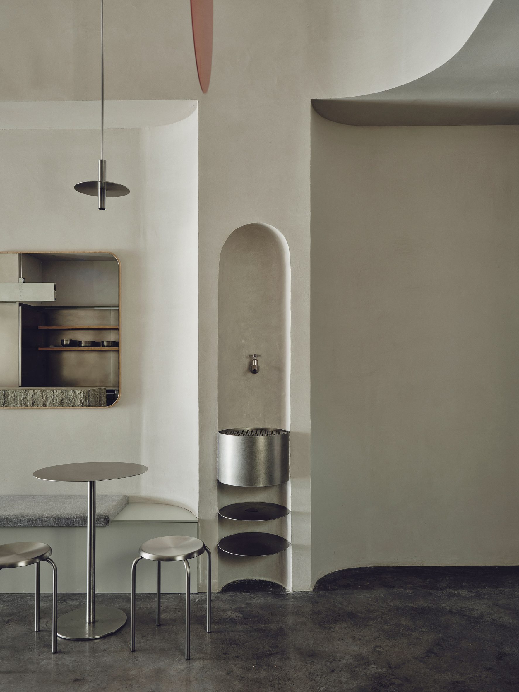 Minimalist cafe seating area with stainless steel tables and stools, bench seating, large rectangular wall mirrors, a custom pendant light and a curved stainless steel basin area set within a wall niche.