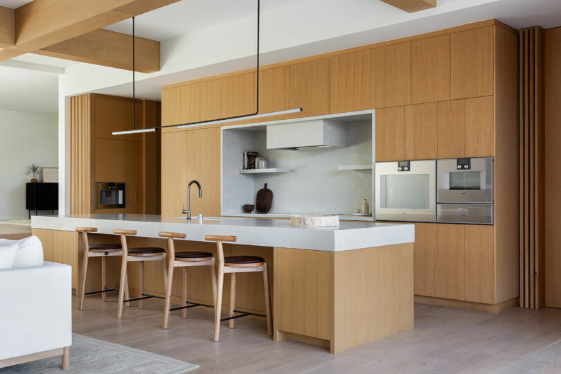 Modern kitchen with light wood cabinetry, white countertops, built-in appliances, a large island with four wooden stools, open shelves, and minimalist decor.