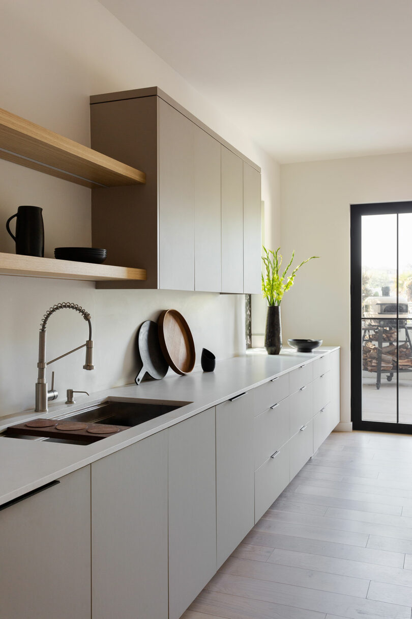 Modern kitchen with light-colored cabinets, open wooden shelves, a stainless steel sink, and a glass door letting in natural light; decorative dishes and a vase with green branches on the counter.