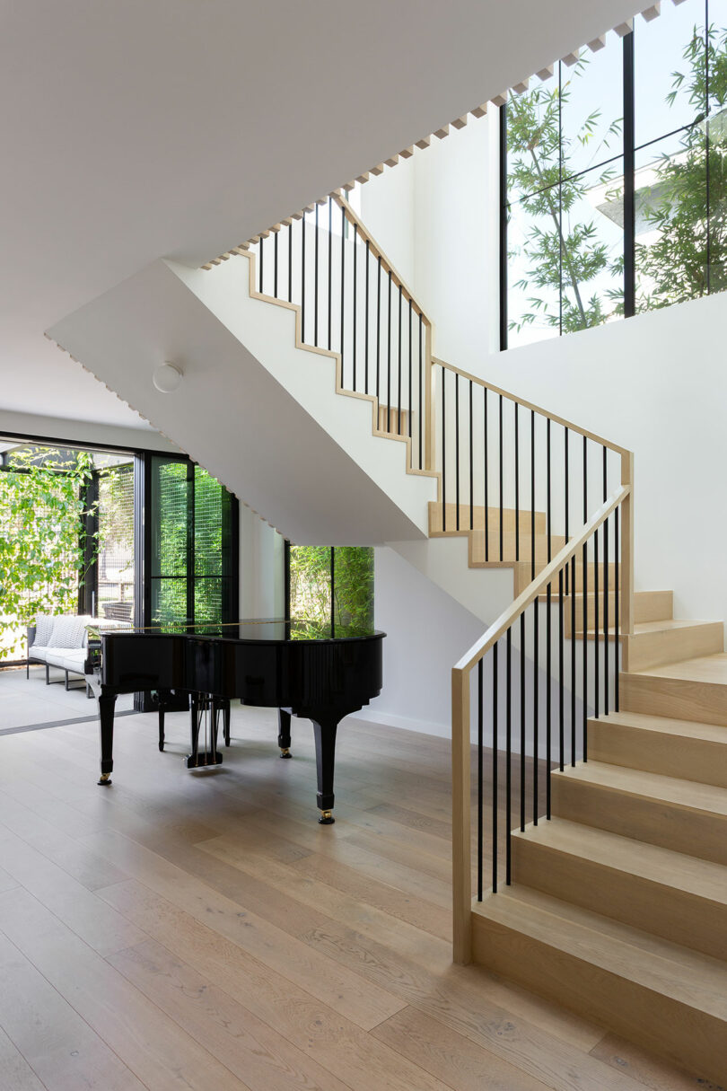 Modern interior with a light wood staircase, black metal railings, and a black grand piano on light wood flooring, large windows showing greenery outside.