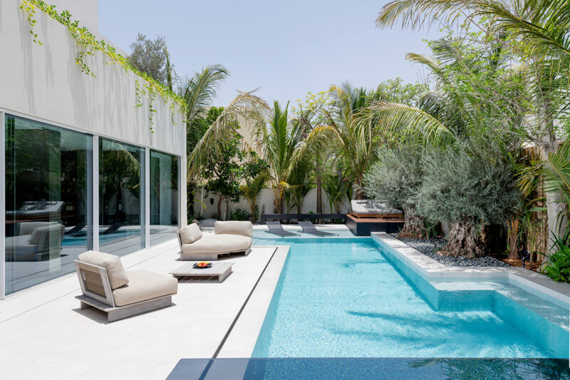 Modern outdoor pool area with white lounge chairs, a clear blue swimming pool, tropical plants, and a glass-walled building on the left.