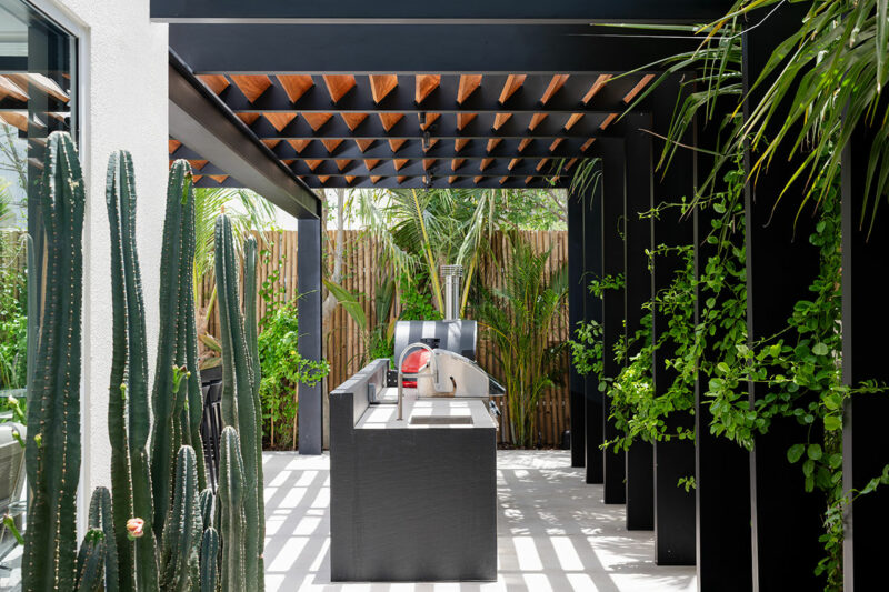 Outdoor kitchen with a modern black pergola, built-in grill, and countertop; surrounded by lush green plants, cacti, and bamboo fencing.