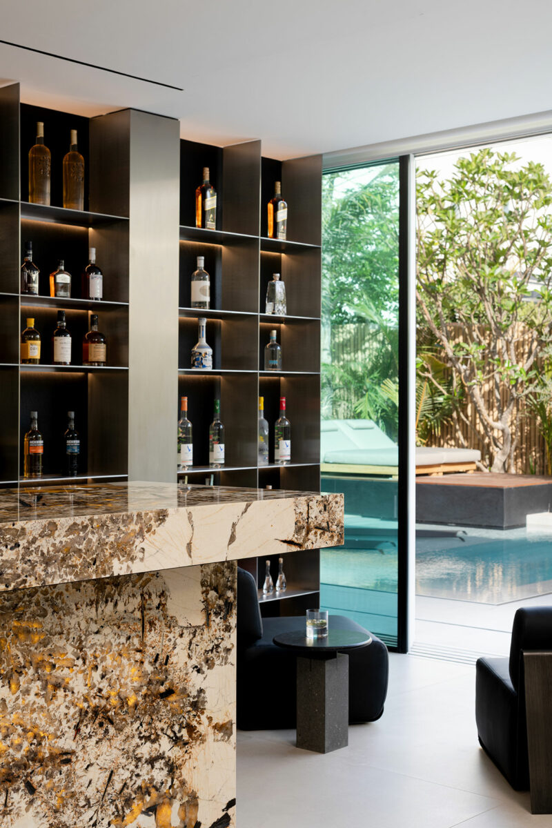 Modern home bar with marble countertop, backlit shelves displaying liquor bottles, black chairs, and a view of a pool and garden through floor-to-ceiling windows.