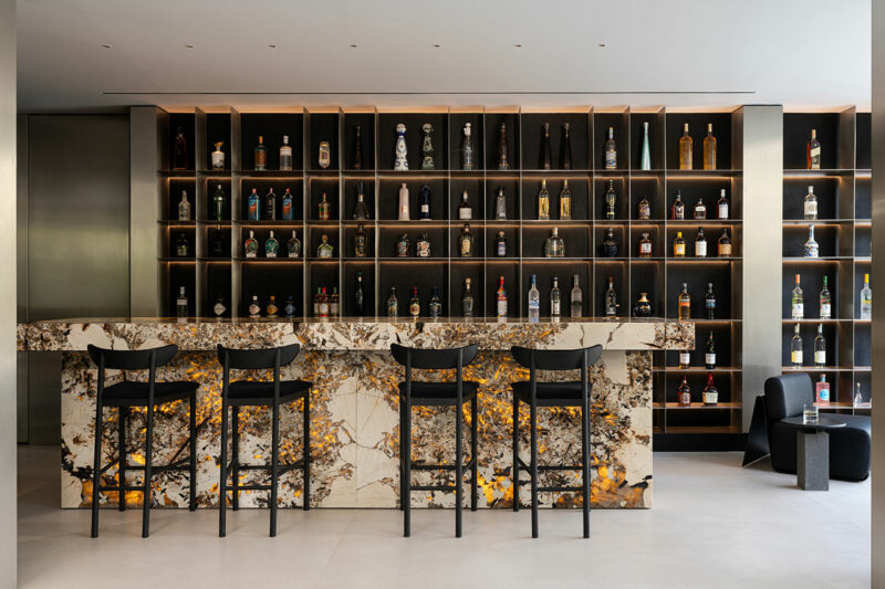 Modern bar with a marble counter, four black barstools, and shelves filled with various liquor bottles against a backlit wall.