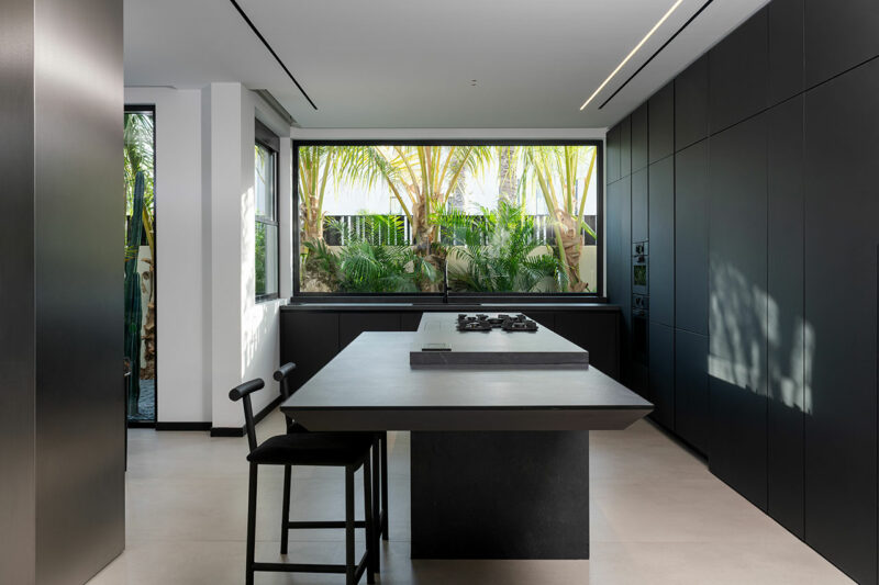 Modern kitchen with matte black cabinetry, a large central island with two stools, and a wide window overlooking lush greenery and palm trees.