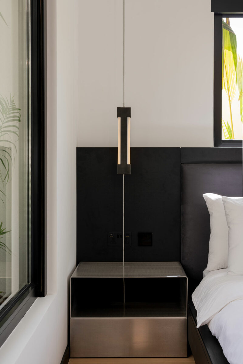 Minimalist bedroom corner with a black headboard, metal nightstand, modern pendant light, and part of a neatly made bed with white linens. A window is visible on the left.