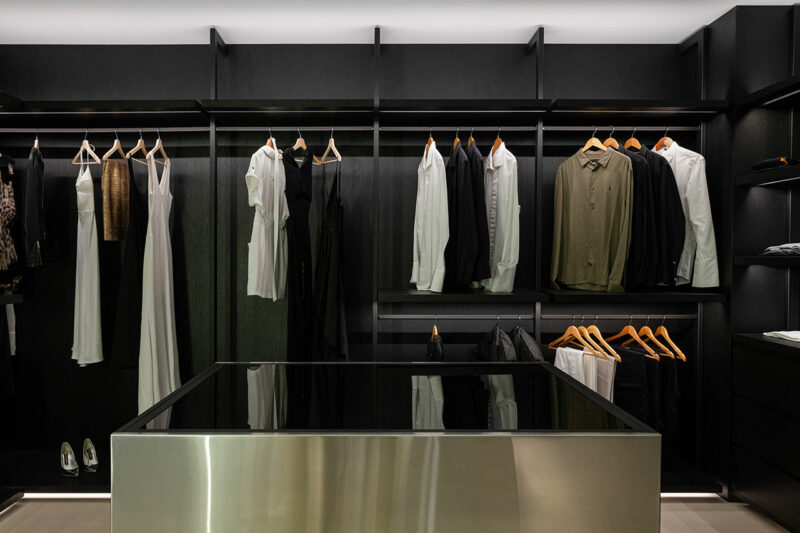 A modern walk-in closet with black shelves and hanging rods, displaying neatly arranged clothes in neutral tones. A reflective island is centered in the room.