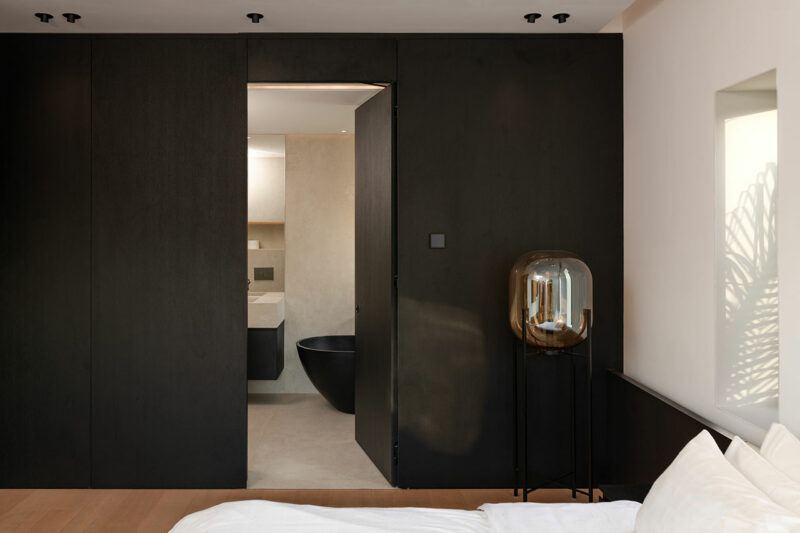 A modern bedroom features a dark wall with a doorway leading to a minimalist bathroom with a black bathtub; a glass lamp stands beside a white bed.