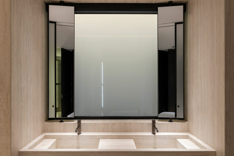 Double sink bathroom vanity with two faucets, a rectangular stone basin, and a large, modern mirror with sliding panels in a neutral-toned setting.