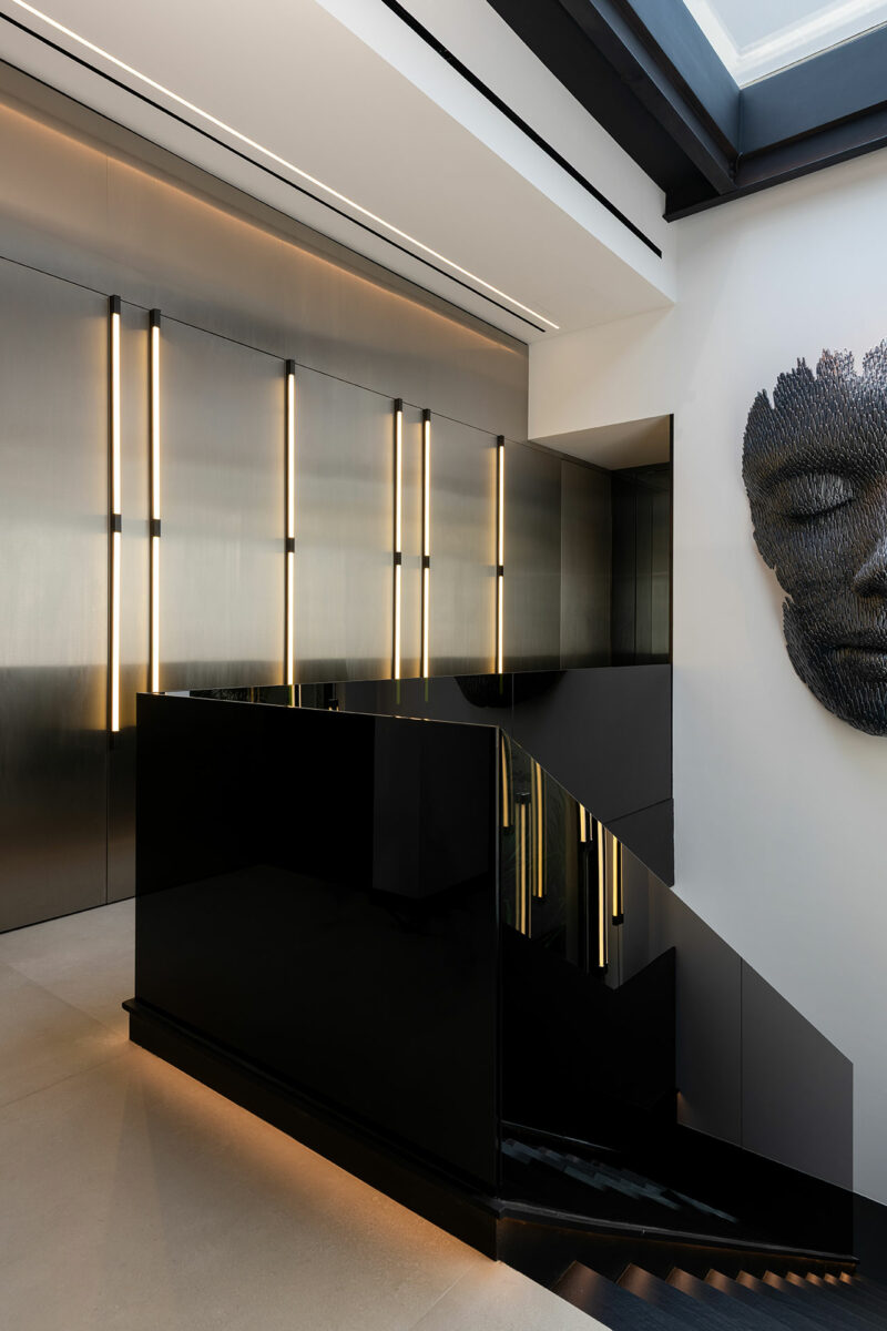Modern interior with a black glossy staircase, vertical wall lights, metallic wall panels, and a textured black face sculpture mounted on a white wall.