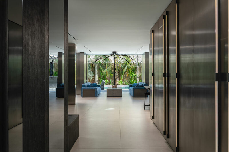 Modern indoor lounge area with blue sofas, large windows, and a view of lush greenery outside. Minimalist design with neutral tones and sleek surfaces.