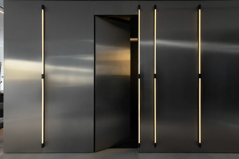 A metallic wall with vertical light strips and a slightly open pivot door in the center.