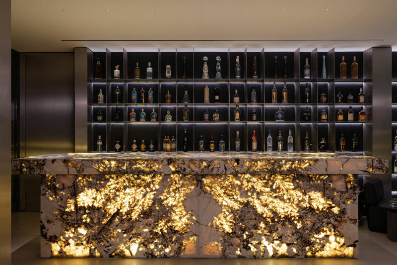 A backlit marble bar counter with illuminated patterns stands in front of shelves lined with assorted liquor bottles in a modern, dimly lit setting.
