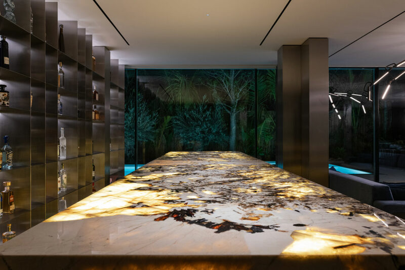 Modern bar area with a backlit marble countertop, glass shelving with bottles, large windows, and greenery visible outside at night.