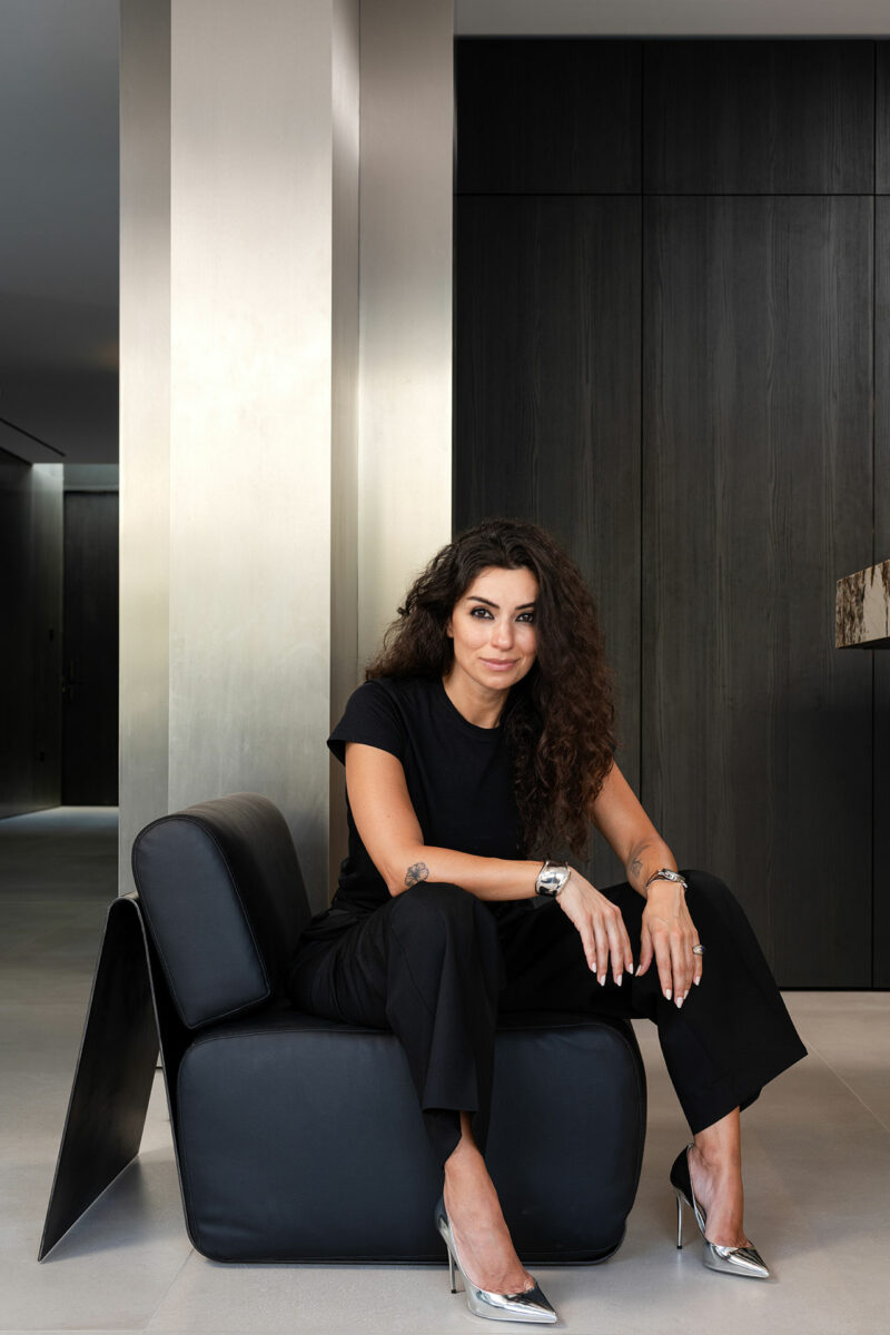 A woman with long curly hair, dressed in black, sits on a modern black chair in a minimalist interior space with dark walls and metallic accents.