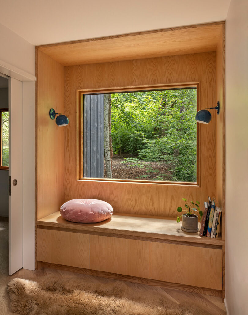A built-in wooden window seat with a pink round cushion, books, a small plant, and two blue wall sconces overlooks a green forest.