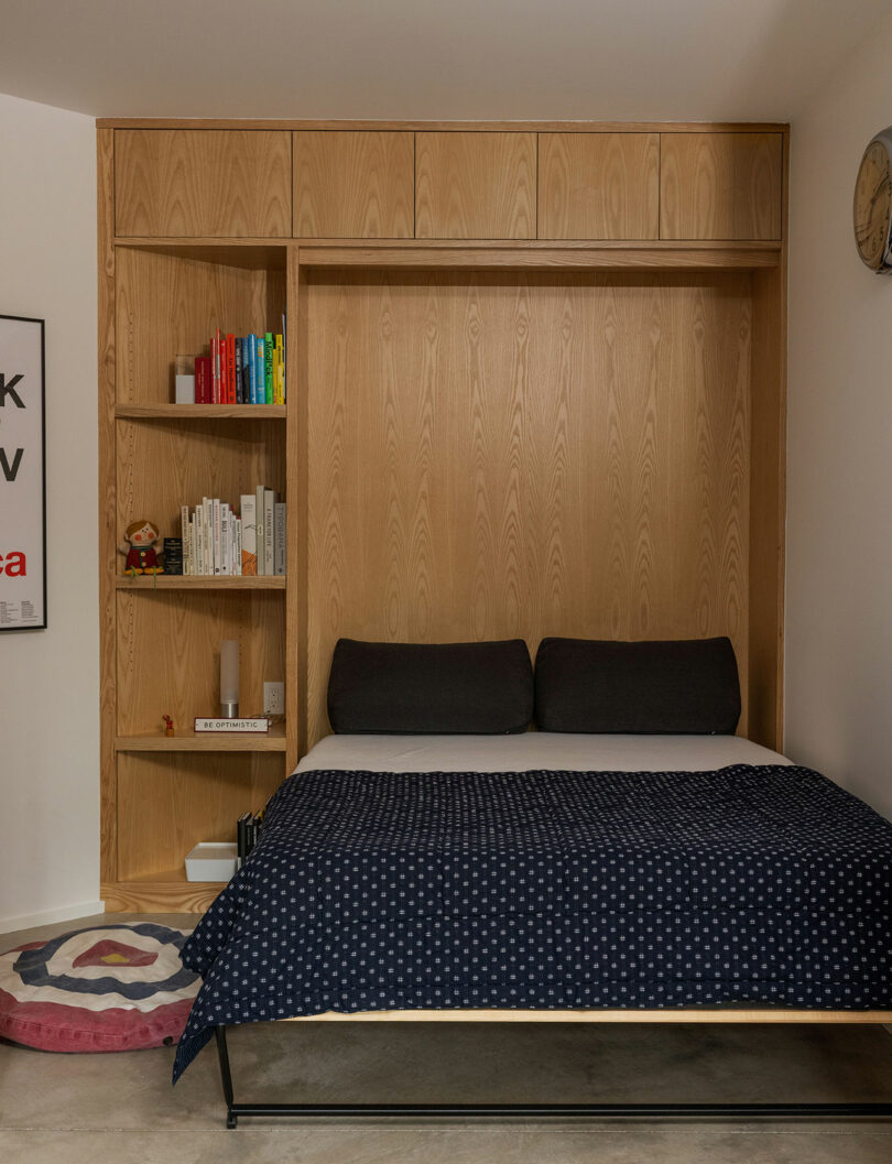 A wooden Murphy bed with a blue polka dot blanket is pulled down next to built-in shelves with books and decorations in a modern room.