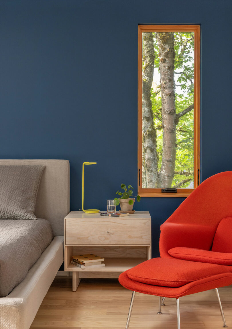 A modern bedroom with a blue wall, beige bed, light wood nightstand, yellow lamp, potted plant, and orange chair by a window showing tree trunks outside.